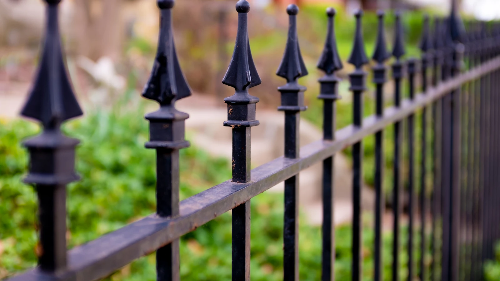 Close-up of a black ornamental metal fence with spear-top pickets beside a garden