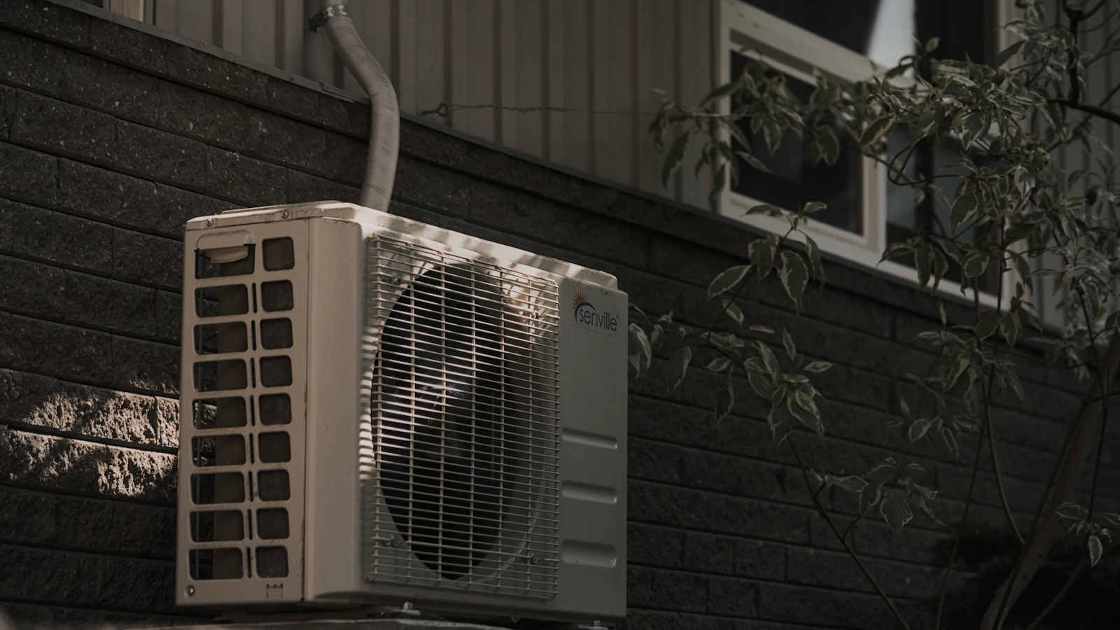 An outdoor air conditioning condenser unit mounted against a dark brick wall