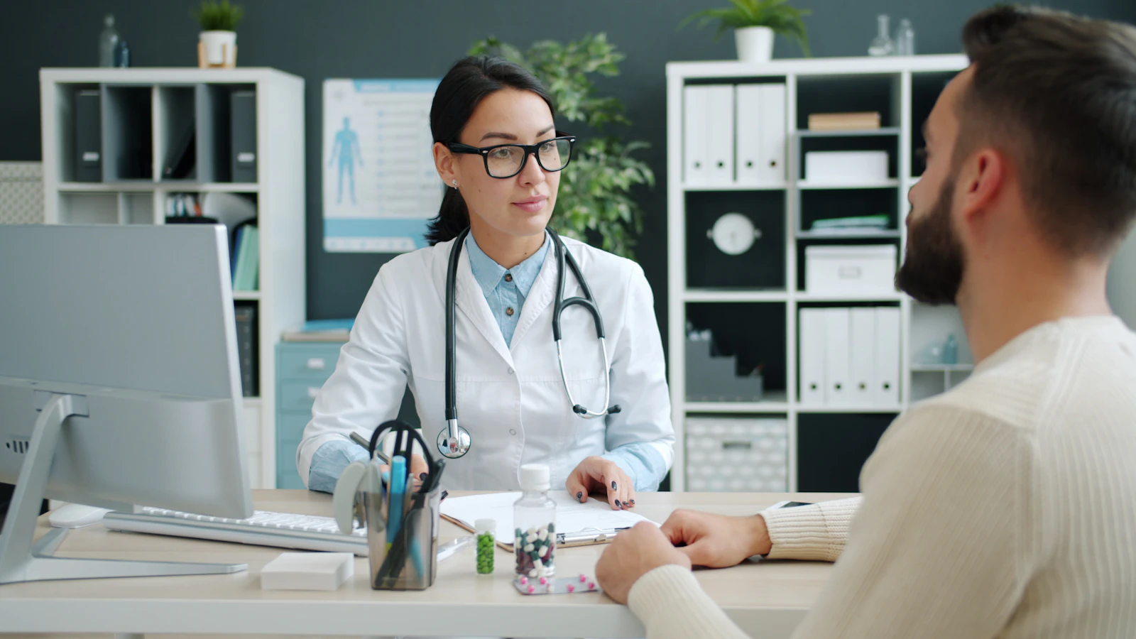 A physician in a white coat talking with a patient across a desk during a consultation